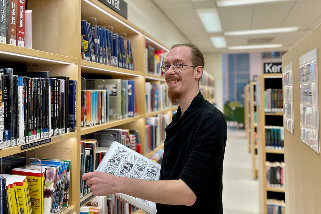 Paavo Rautiainen Lahden kirjaston hyllyjen edessä selaamassa sarjakuvateosta